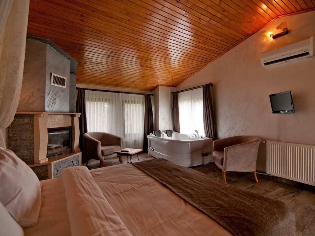 A bedroom with a wooden ceiling, a fireplace, a large bed, a sitting area with chairs, a bathtub, and a television mounted on the wall.