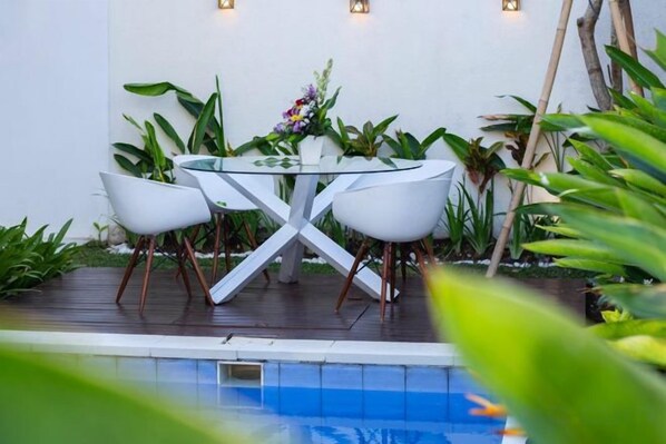 A glass-top table with four chairs, surrounded by plants and a vase of flowers, set on a wooden deck by a pool.