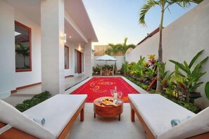 Un patio con alfombra roja, sillas de descanso, una mesa de madera con una botella y plantas tropicales.