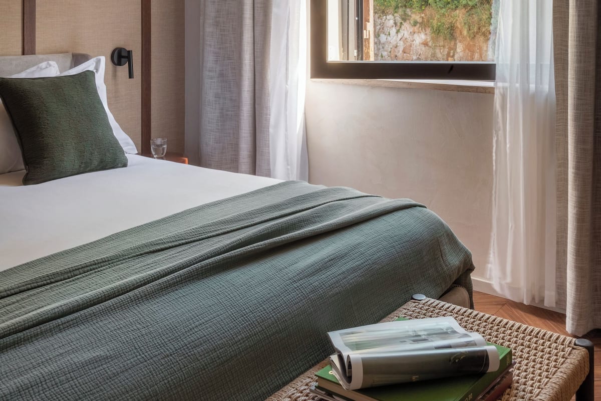 A hotel room with a bed, a window with a view of greenery, a bedside table with books, and a wall-mounted light.