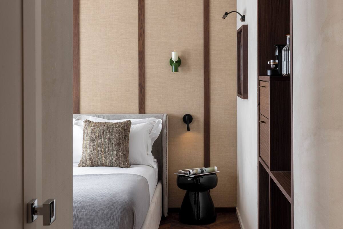 A narrow hotel room with a bed, a small bedside table, a wall-mounted light, and a wooden wardrobe.