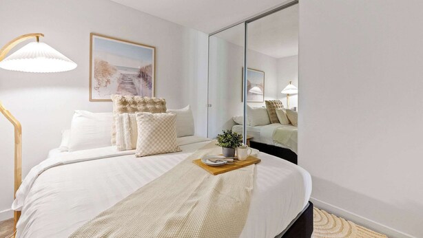 Een keurig opgemaakte slaapkamer met een bed, bedlampje en een spiegel die de kamer weerspiegelt.