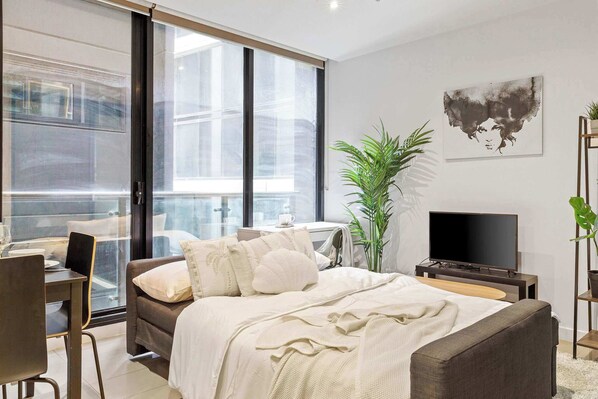 A modern bedroom with a large bed, a desk, a chair, a TV, and a plant.