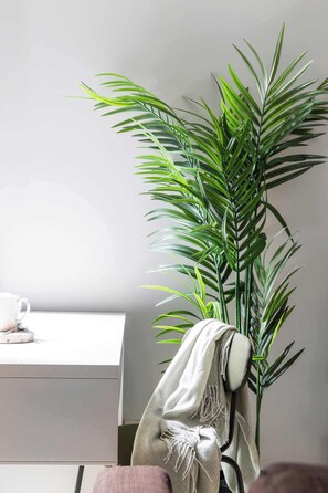 Una habitación con una planta grande en maceta, un escritorio blanco con una taza y un plato, y una silla con una manta.