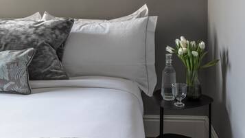 A bedroom with a bed, pillows, a bedside table with a vase of flowers, and a small chair.