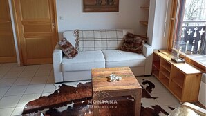 Een woonkamer met een witte bank, een houten salontafel, een kussen van runderhuid, een houten deur, een bergschildering aan de muur, en een raam met uitzicht op bomen.