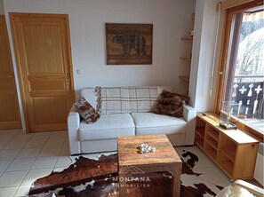 Un salon avec un canapé blanc, une table basse en bois, un tapis en peau de vache, une porte en bois, une sculpture d’ours fixée au mur et une fenêtre donnant sur des arbres.