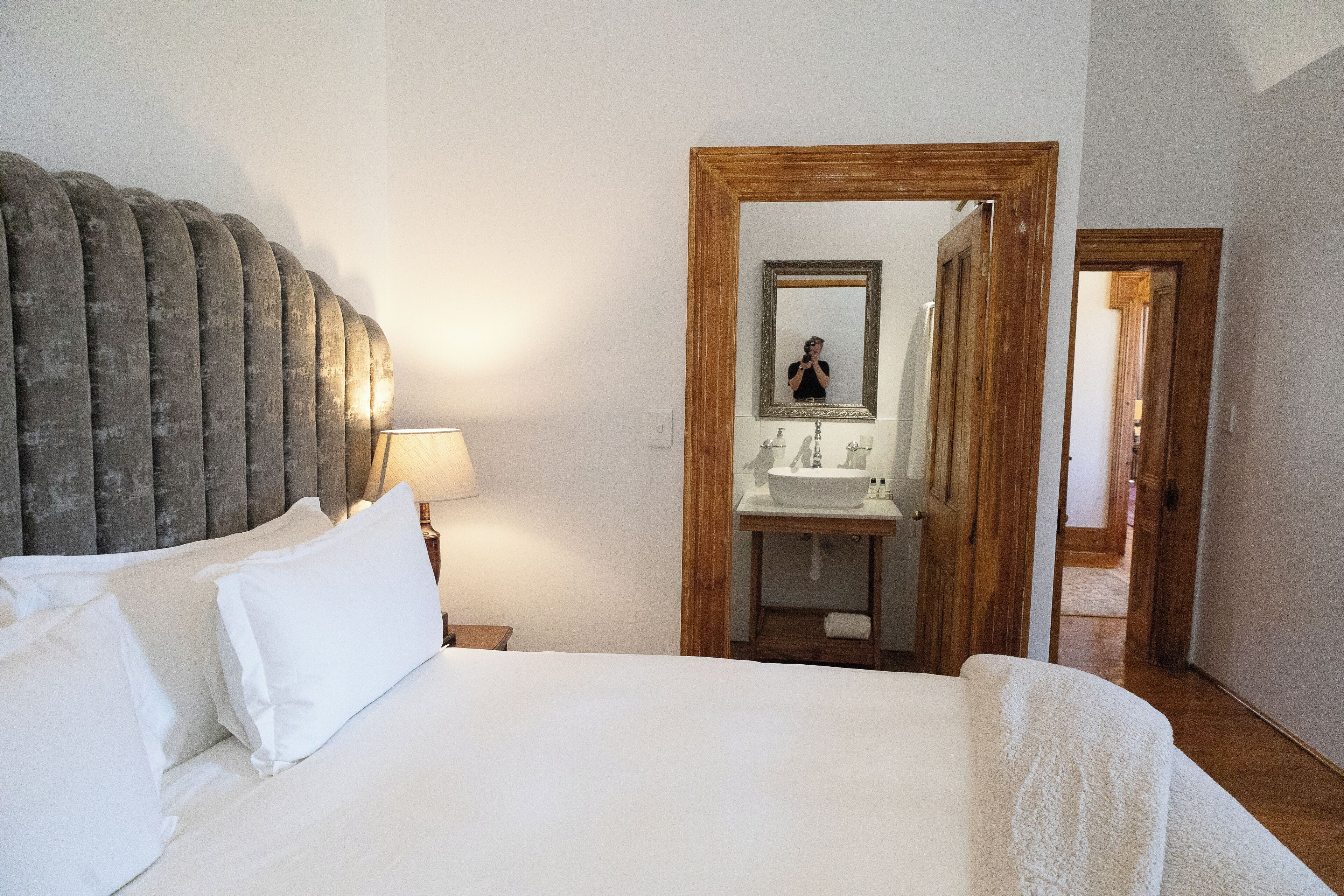 A hotel room with a large bed, a wooden headboard, a bedside table with a lamp, a bathroom with a sink and mirror, and a wooden door leading to another room.