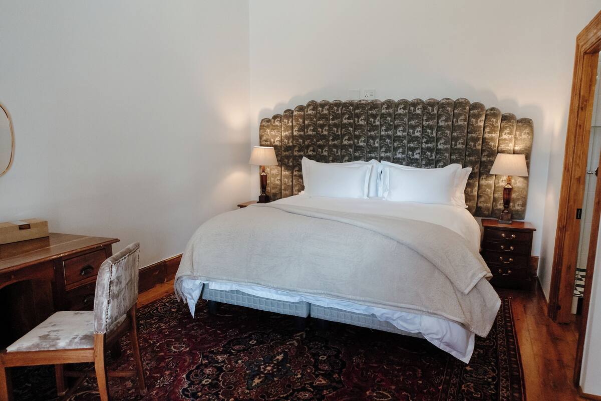 Ein ordentlich bezogenes Bett mit Kopfteil, ein Nachttisch aus Holz mit einer Lampe, ein Stuhl und ein gemustertes Teppich.