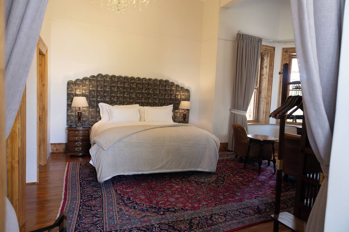 Ein Schlafzimmer mit einem großen Bett, einem hölzernen Kopfteil, einem roten Teppich mit Muster sowie einem kleinen Tisch und zwei Stühlen.