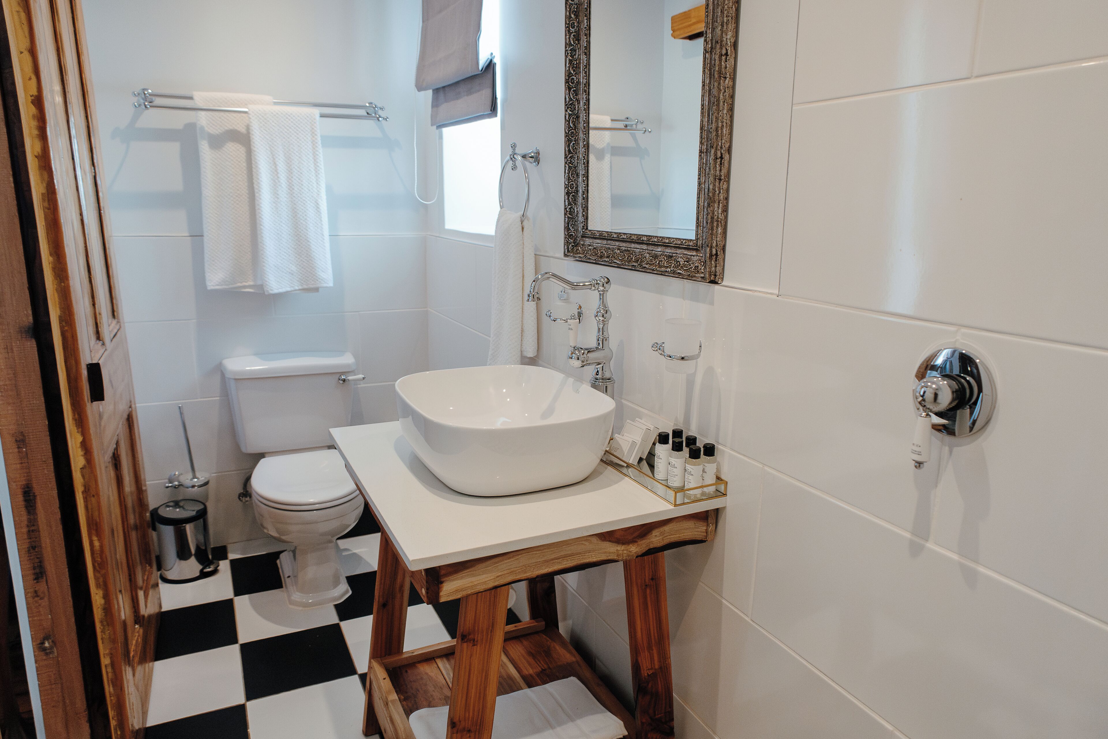 Un baño con lavamanos, inodoro y tocador blancos. Hay un taburete de madera debajo del tocador, y se ve un cubo de basura.