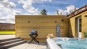 A hot tub in a backyard with a wooden wall, steps leading up to it, and a grill on a cart.