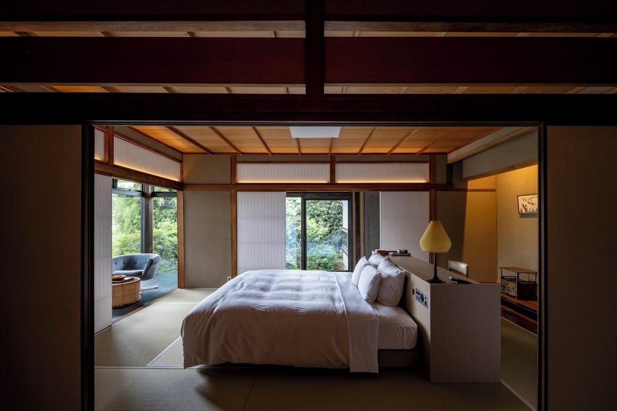 A traditional Japanese-style bedroom with tatami flooring, a bed with white bedding, a desk with a lamp, and a view of greenery through large windows.