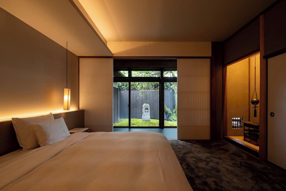 A modern hotel room with a large bed, a bedside table, a nightstand, a wooden bench, and a sliding door leading to a garden with a stone lantern.