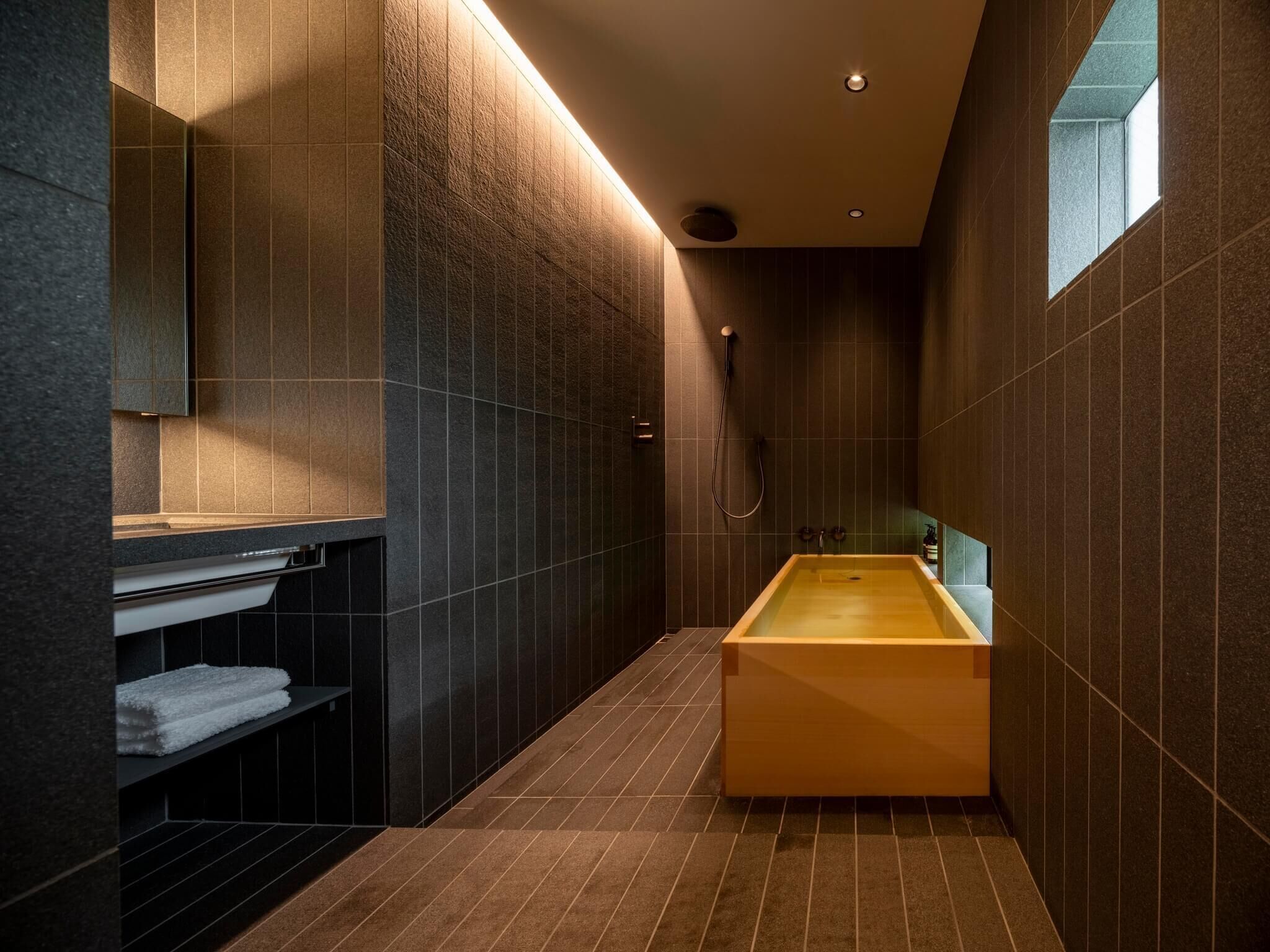 A modern bathroom with a wooden bathtub, dark tiled walls, and a built-in shelf with towels.