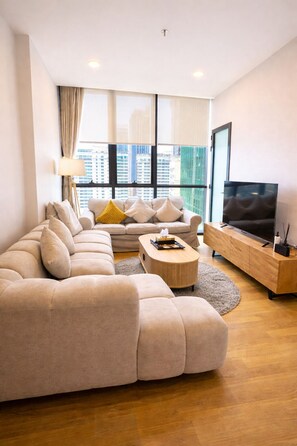 A modern living room with a sectional sofa, a coffee table, a TV on a wooden stand, and large windows with a city view.