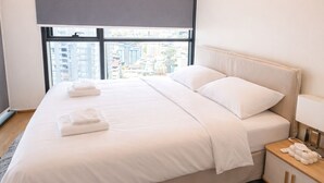 Quarto de hotel moderno com uma cama grande, uma mesinha de cabeceira com luminária e vista para a cidade pela janela.