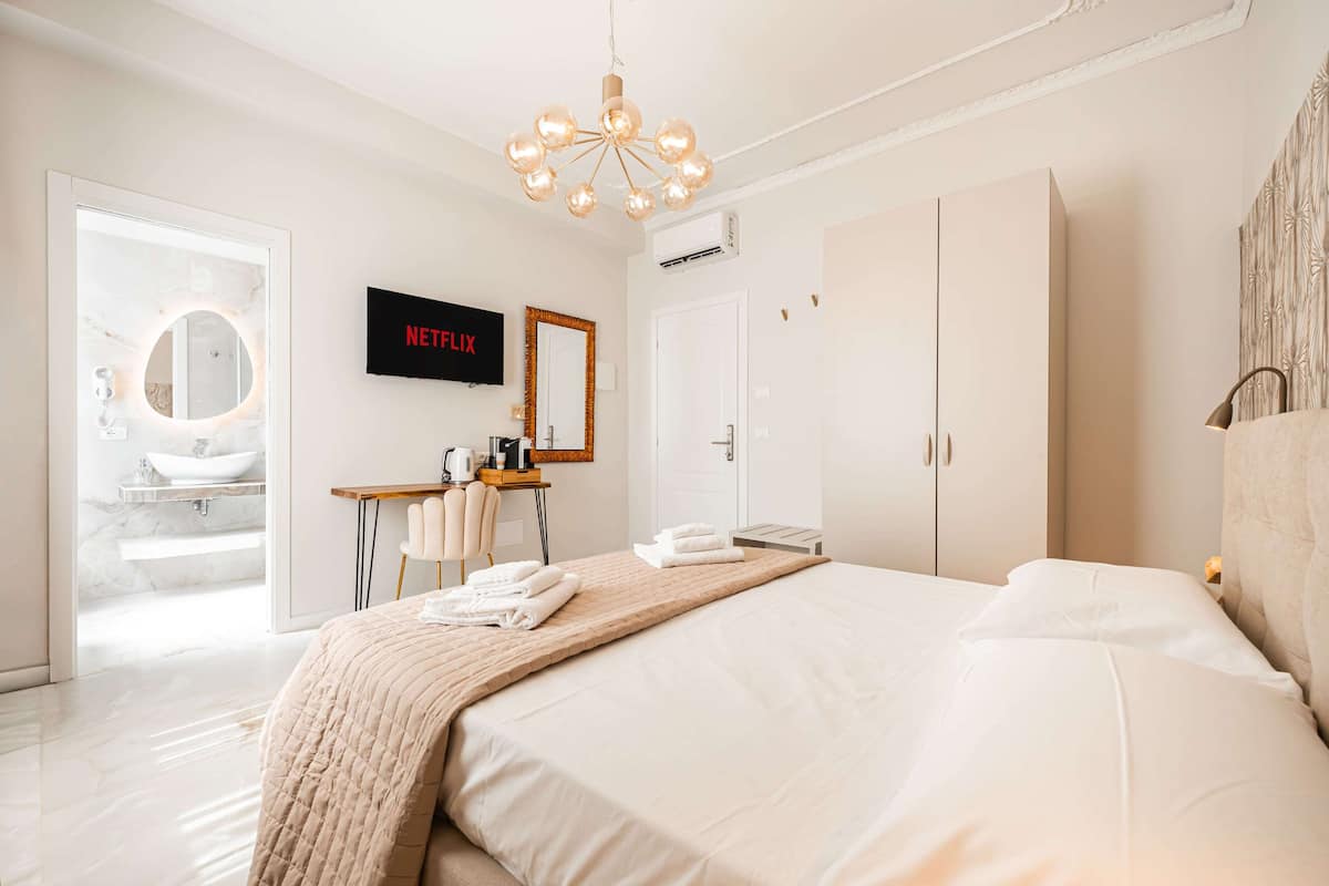 Moderni hotellihuone, jossa on suuri sänky, työpöytä tuolilla, televisio sekä kylpyhuone, jossa on peili ja pesuallas.