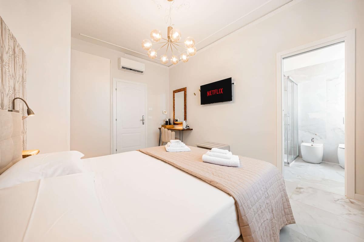 Hotellihuone, jossa on sänky, yöpöytä, peili, seinälle asennettu televisio ja kylpyhuone, joka näkyy avoimesta ovesta.