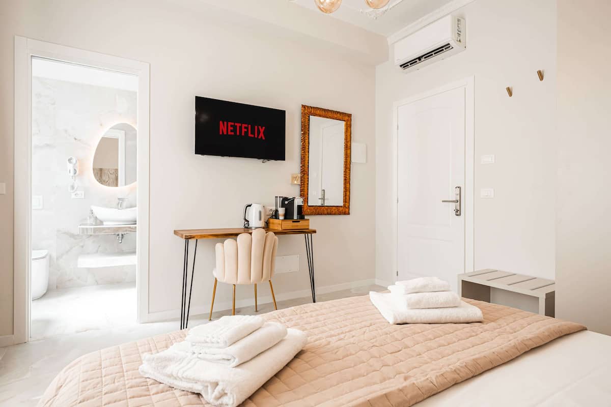Moderni hotellihuone, jossa on sänky, työpöytä tuolilla, televisio, jossa näkyy Netflix, kylpyhuone, jossa on wc ja pesuallas, sekä peili.