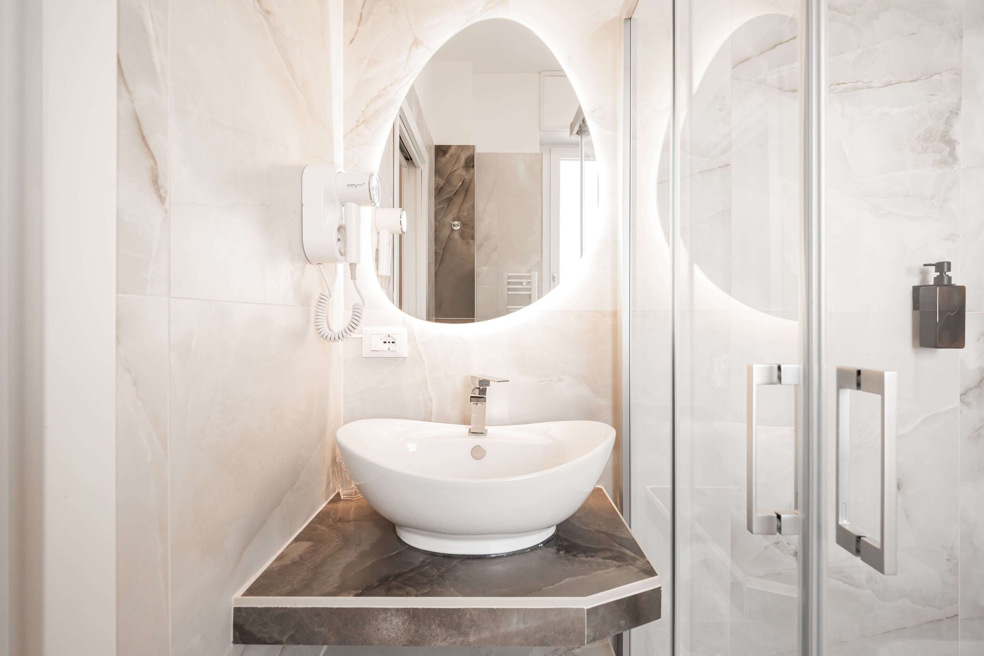 Un baño moderno con un espejo ovalado grande, un lavamanos blanco tipo recipiente y una pared revestida de mármol.
