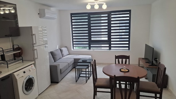 A compact living space with a kitchenette, a gray sofa, a dining table with chairs, and a large window with blinds.
