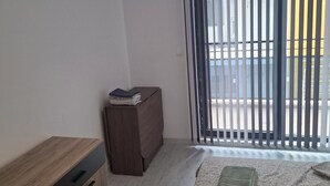 A room with a wooden desk, a small cabinet, a bed with a patterned bedspread, and a window with vertical blinds.