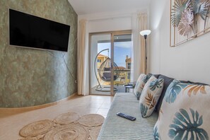 A living room with a flat-screen TV, a sofa with floral cushions, a woven rug, and a view of a balcony with a hanging chair.