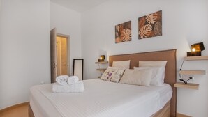 Una cama bien hecha con sábanas blancas y una toalla enrollada, un cabecero de madera, una mesita de noche con una lámpara y dos obras de arte colgadas en la pared.