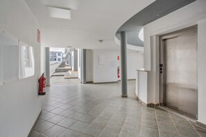 Un couloir au sol carrelé, un extincteur, un ascenseur fermé et un escalier en arrière-plan.