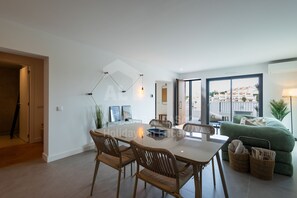 A modern living room with a dining area, featuring a large table, wicker chairs, a green sofa, and a view through large windows.