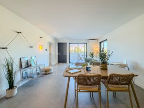 A spacious living area with a dining table, two wicker chairs, a potted plant, and a view through a glass door.