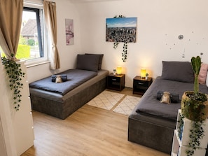 Ein Schlafzimmer mit zwei Betten, einem Fenster mit Vorhängen, einem Stadtbild und einer Kakteenpflanze.