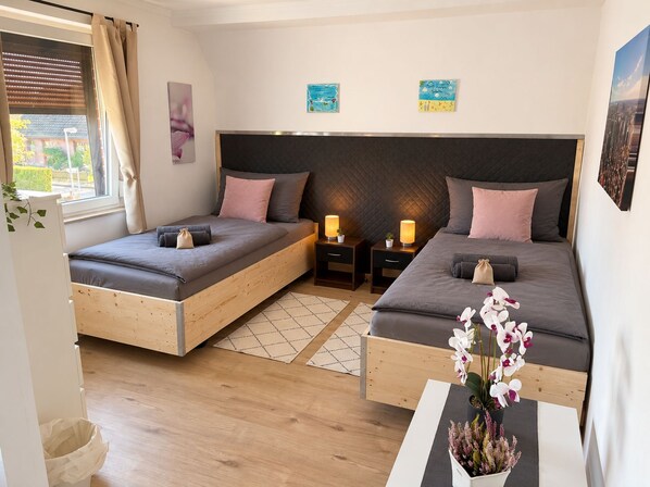 A bedroom with two beds, a window with blinds, a bedside table with lamps, and artwork on the wall.