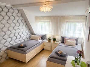 A modern bedroom with two beds, a wooden headboard, a small table with a lamp, and a window with sheer curtains.