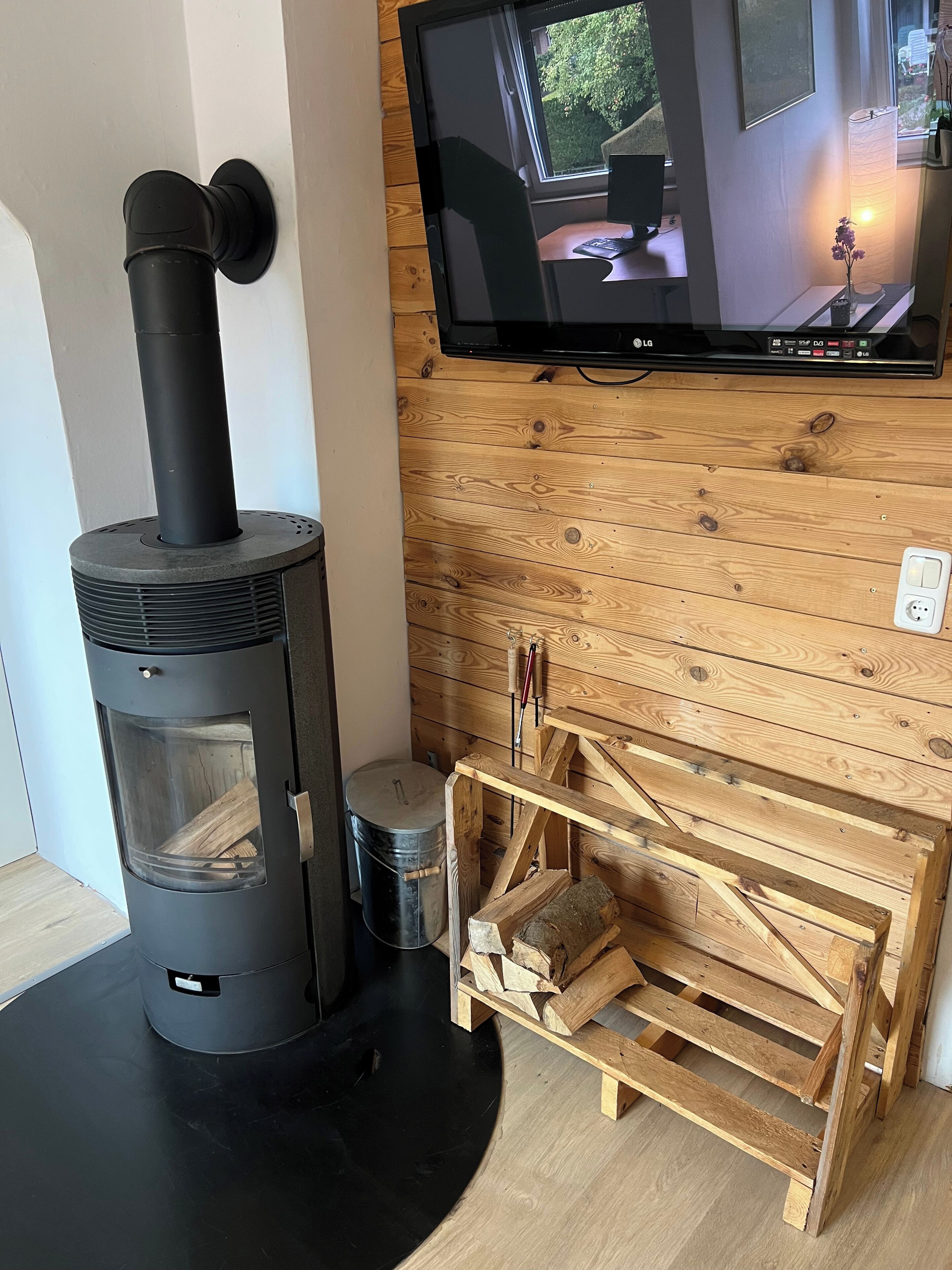 A wood-burning stove, a television mounted on a wooden wall, and a small wooden table with a chair.