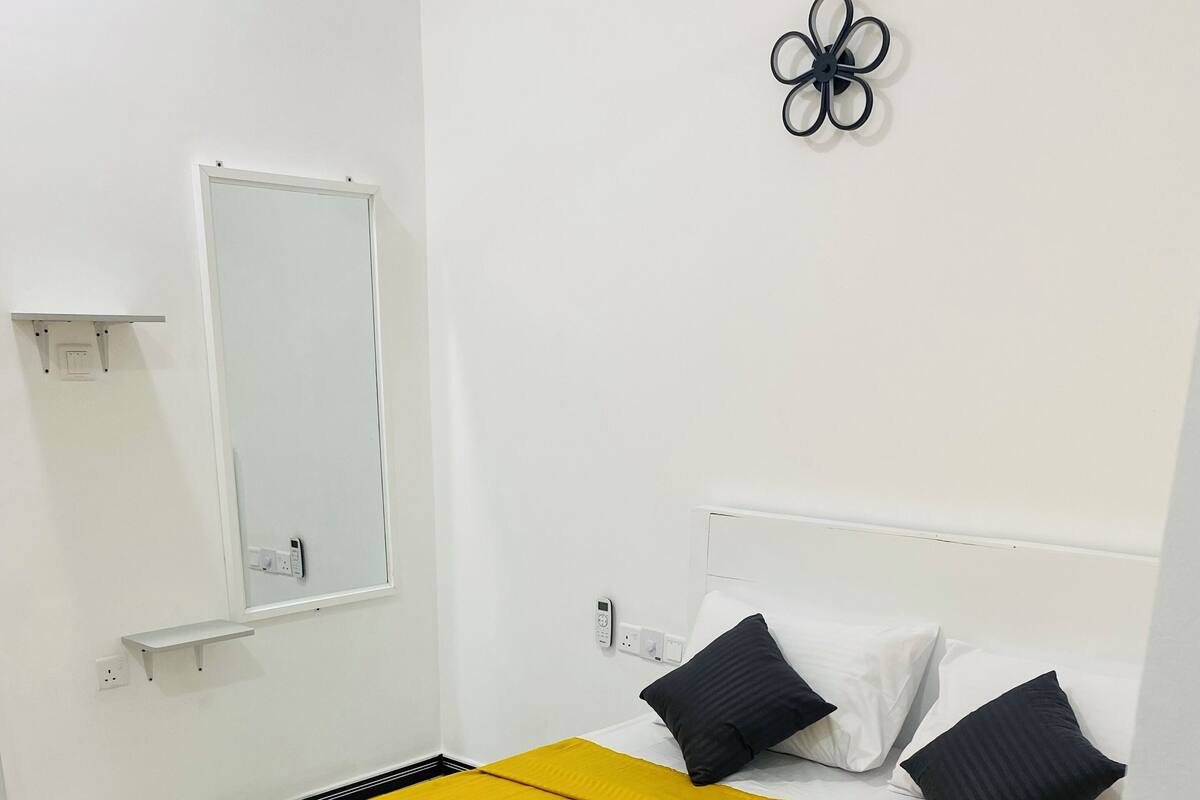 A hotel room with a bed, white walls, a mirror, a small shelf, and a wall-mounted light fixture.