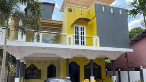 A two-story house with a balcony, a car parked in front, and a small table with chairs outside.