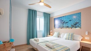 Habitación de hotel con dos camas, ventilador de techo, un ventanal con cortinas y un cuadro en la pared.