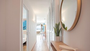 Un pasillo con suelo de madera, una puerta blanca, un espejo y una planta sobre un estante de madera.