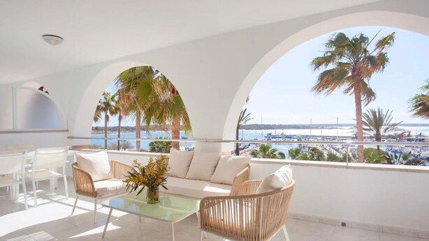 Un balcón iluminado por el sol con un sofá, sillas y una mesa, con vistas a un puerto deportivo y palmeras.