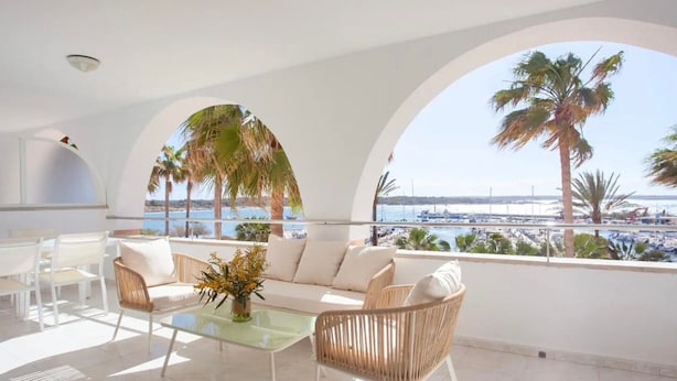 A sunlit balcony with a sofa, chairs, and a table, overlooking a marina and palm trees.