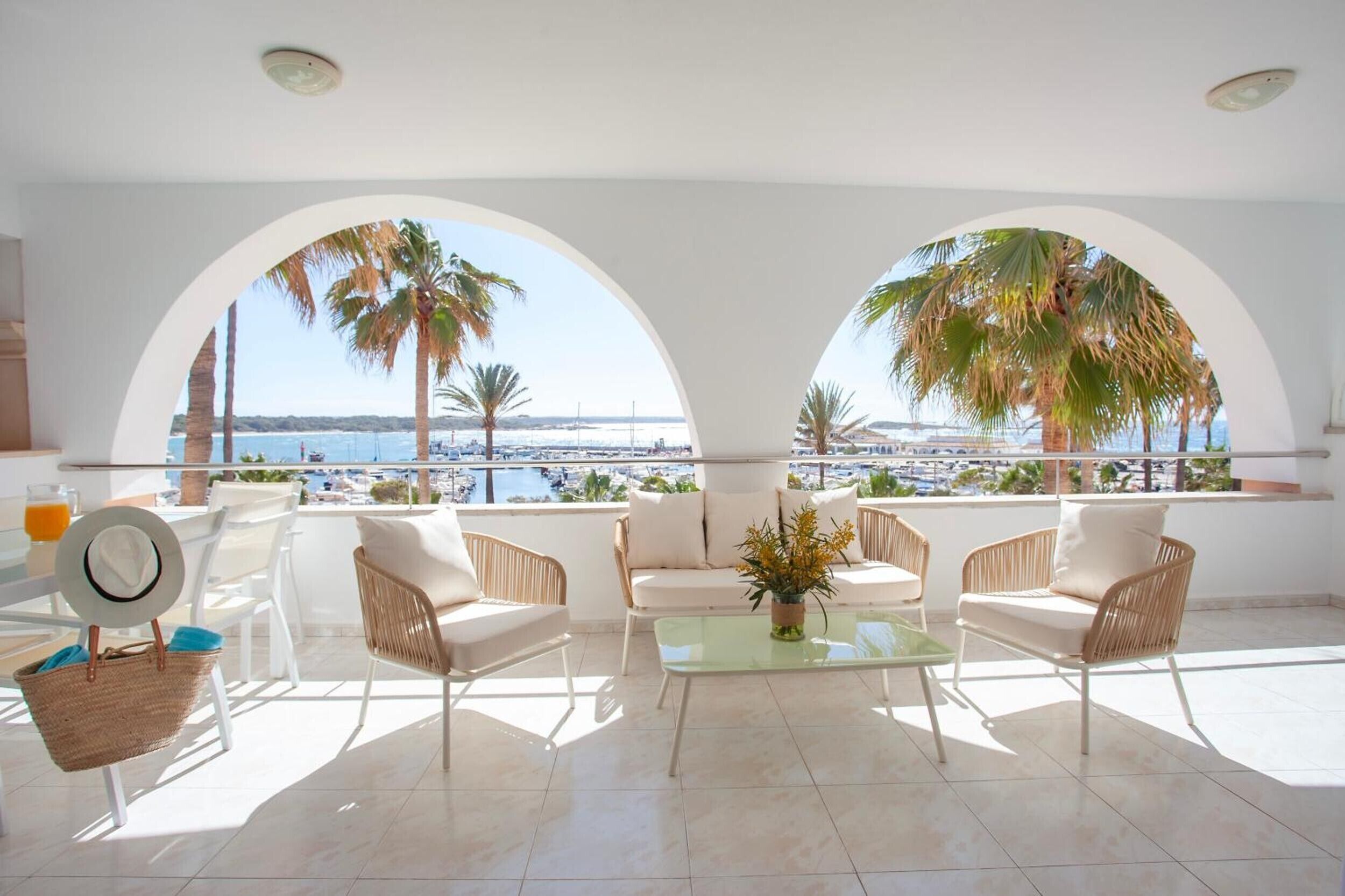 A sunlit room with white furniture, a glass table, and a view of the sea through arched windows.