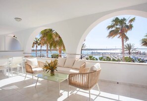 A sunlit balcony with a sofa, chairs, and a table, overlooking a marina and palm trees.
