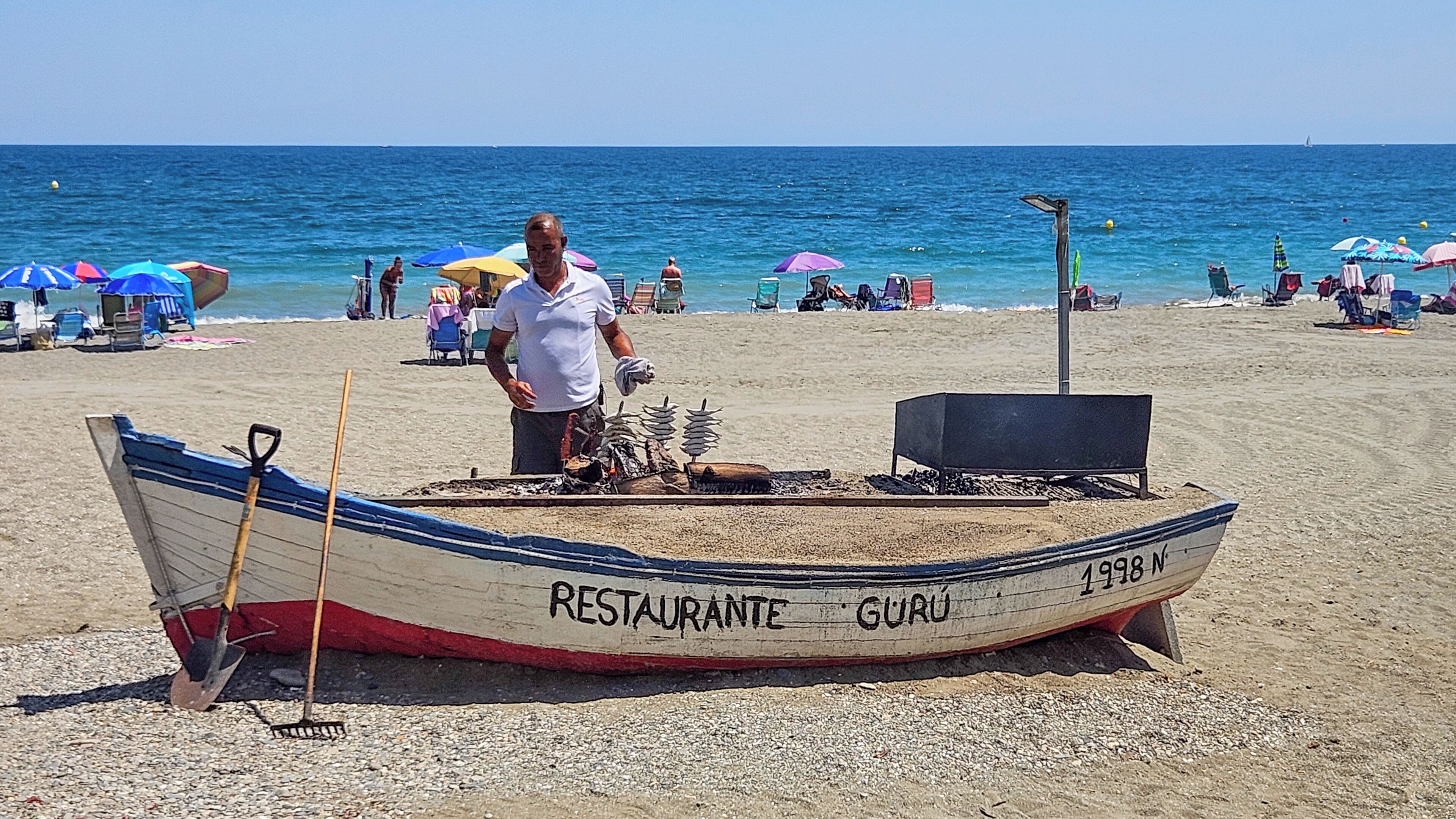 A boat with 'RESTAURANTE GURÚ' written on it, parked on a sandy beach with umbrellas and people in the background.