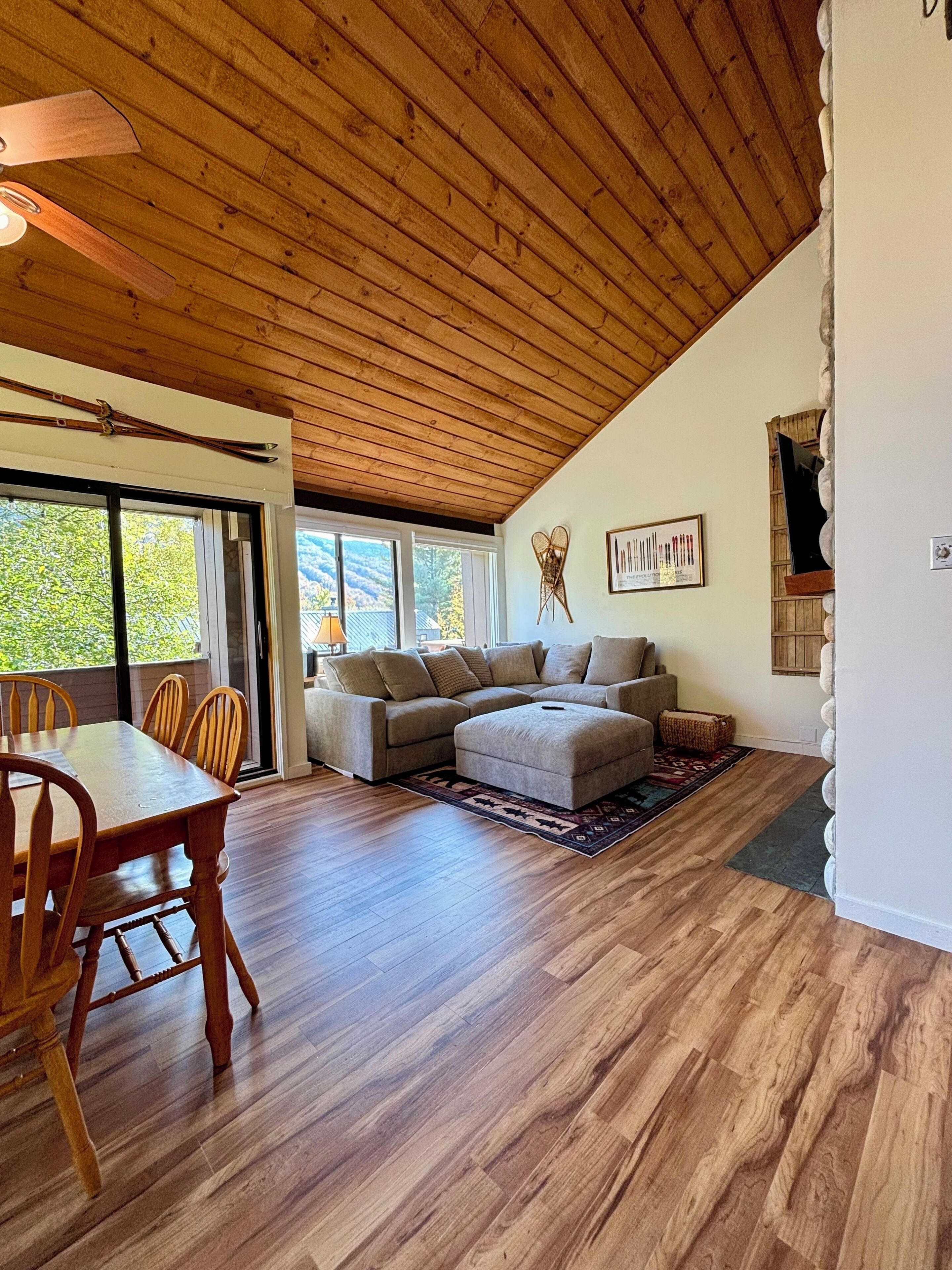 A spacious living area with a wooden ceiling, a large sofa, a dining table with chairs, and a view of greenery outside.