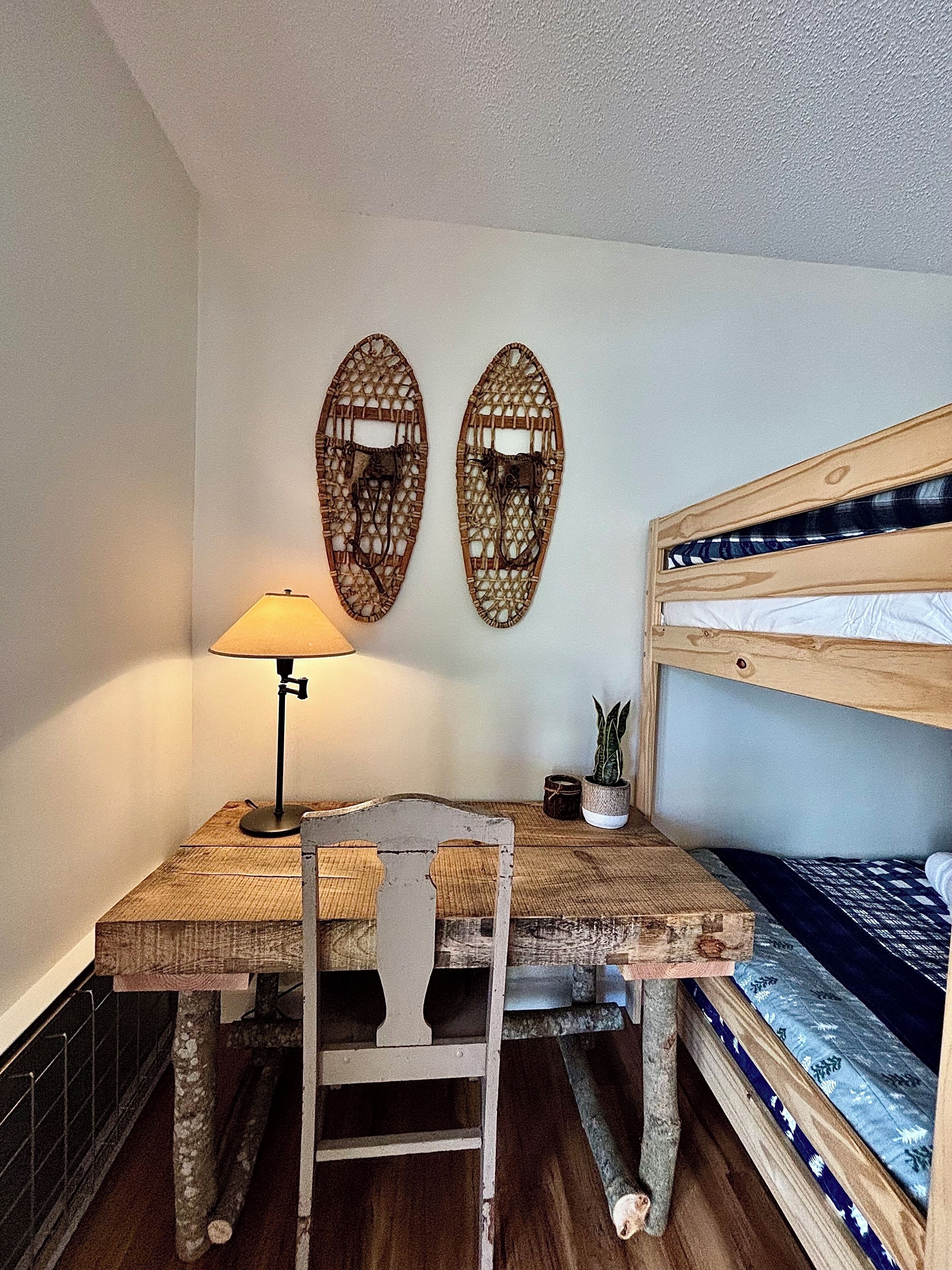 Een kleine kamer met een houten bureau, een stoel, een lamp, een stapelbed en twee decoratieve wandpanelen.