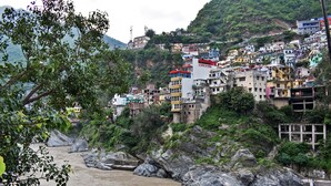 A hillside settlement with colorful buildings, a river in the foreground, and a rocky terrain. - Hotel US Tower (Devaprayāg)
