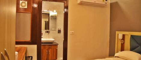 A hotel room with a bed, a wooden wardrobe, a bathroom with a sink and mirror, and an air conditioning unit.