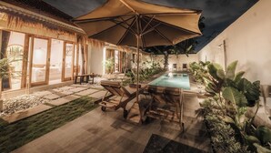 A well-lit outdoor pool area with lounge chairs and a large umbrella, surrounded by potted plants and a thatched-roof structure. - Hiraya Uluwatu (Pecatu)